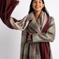 Model wearing two Essential Travel Wraps around her neck.  One is the brick colorway and one is the farro colorway.  She is wearing a plaid coat and smiling.  One side of the wraps has been thrown in the air.